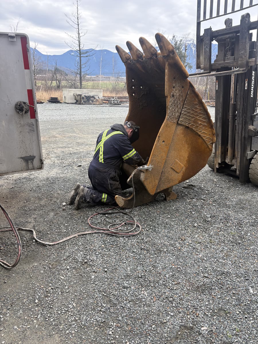 Mazdock Mechanical technician welding an excavator bucket in hi-vis PPE with Agassiz BC mountains behind